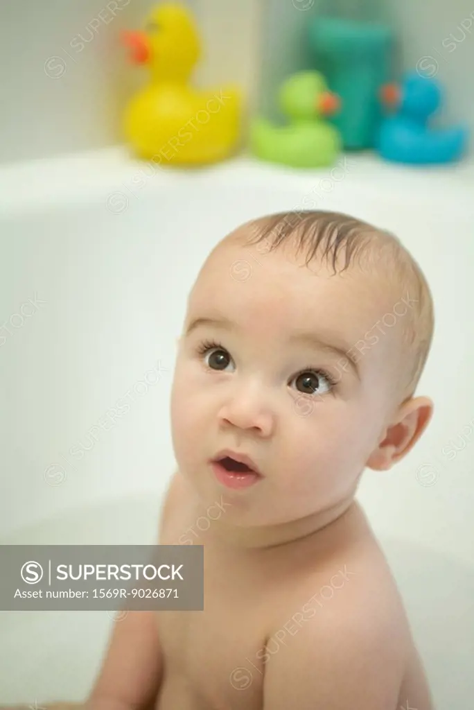 Naked baby sitting in bathtub, looking up, wide eyes