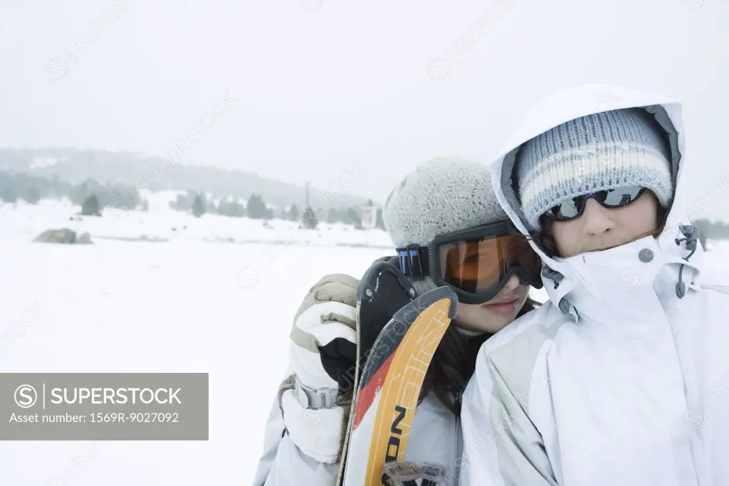 Two young skiers standing side by side, looking at camera, one's head on the other's shoulder