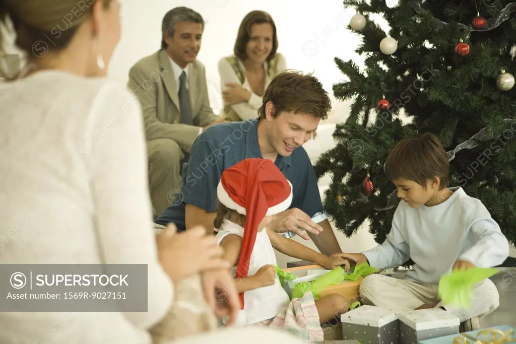 Father and two children sitting by Christmas tree, opening presents together, family watching