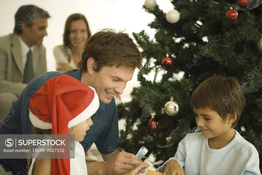 Father and two children sitting by Christmas tree, opening presents together, daughter wearing Santa hat