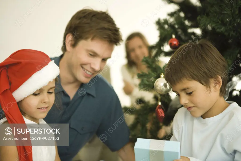Father and two children sitting by Christmas tree, daughter wearing Santa hat, son opening present