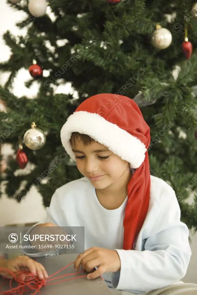 Boy opening present in front of Christmas tree, wearing Santa hat, smiling