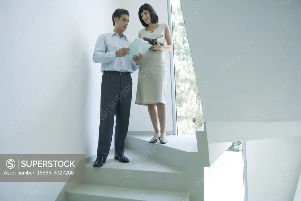 Mature businessman speaking to young female colleague on stairs, showing document