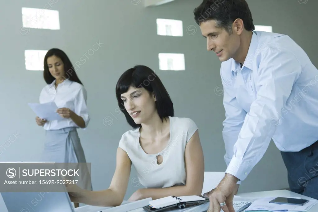 Young businesswoman working at computer, speaking to male colleague standing next to her