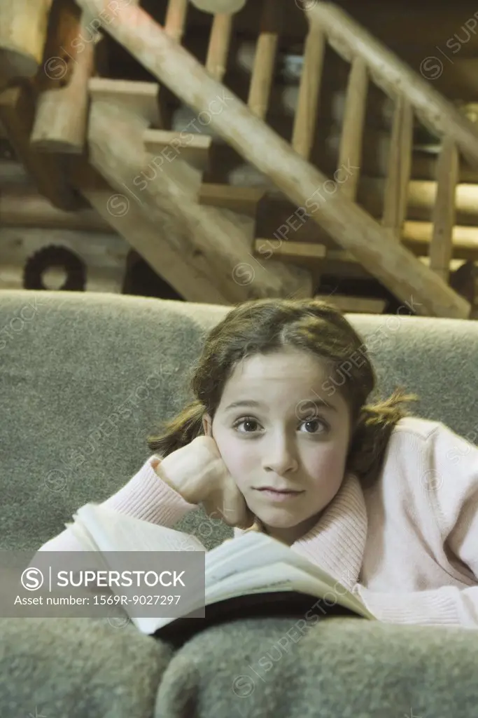 Preteen girl lying on sofa with book, looking at camera