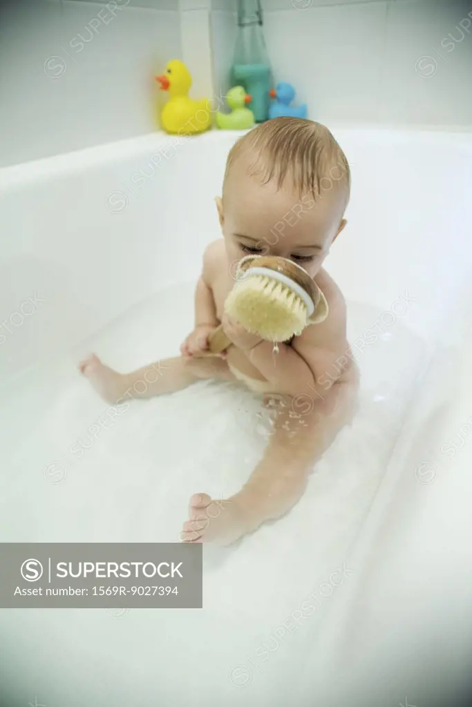 Baby taking bath, holding bath brush against face, full length