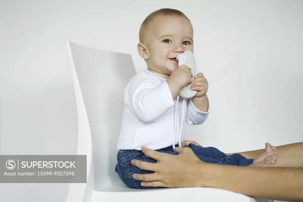 Baby sitting in chair putting shoe in mouth, mother's arms holding baby's legs