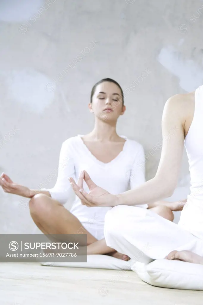 Two young women sitting in lotus position, one's eyes closed, cropped view