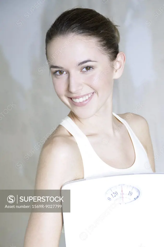 Young woman holding bathroom scale, smiling at camera, portrait