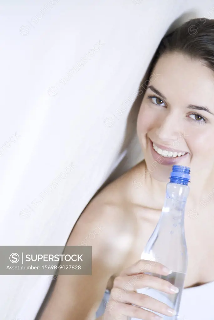 Young woman leaning against wall, holding water bottle, smiling at camera