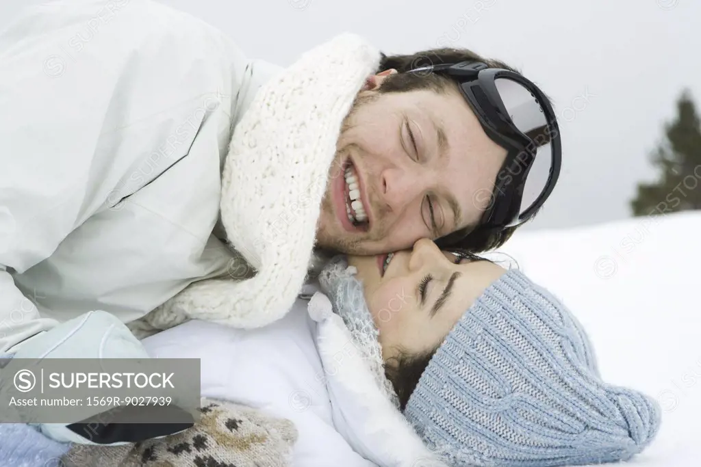 Young couple reclining in snow together, smiling, eyes closed, one on top of the other