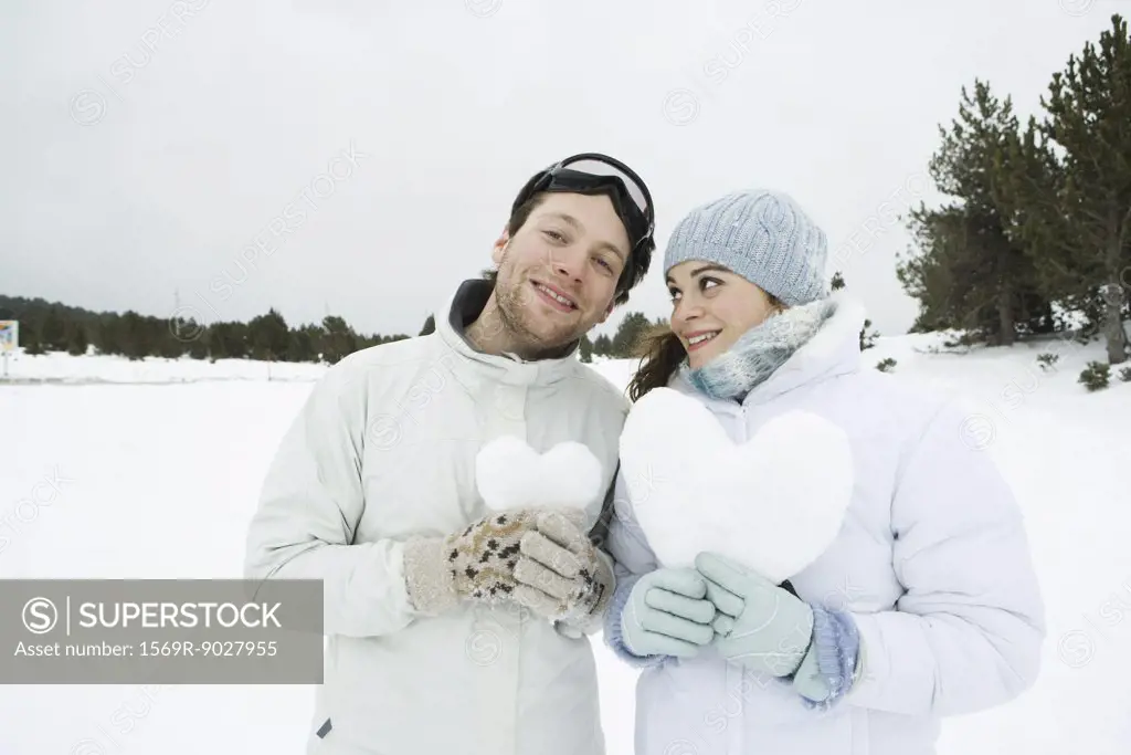 Young couple standing side by side, holding hearts made of snow, portrait