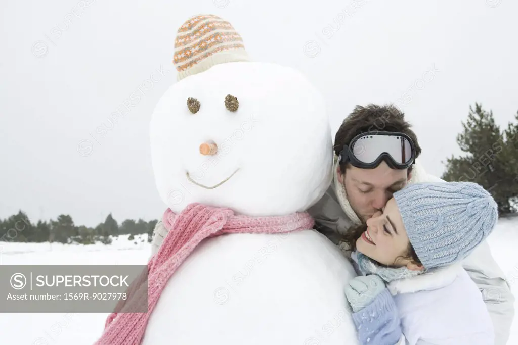Young couple next to snowman, man kissing woman on cheek, eyes closed, portrait
