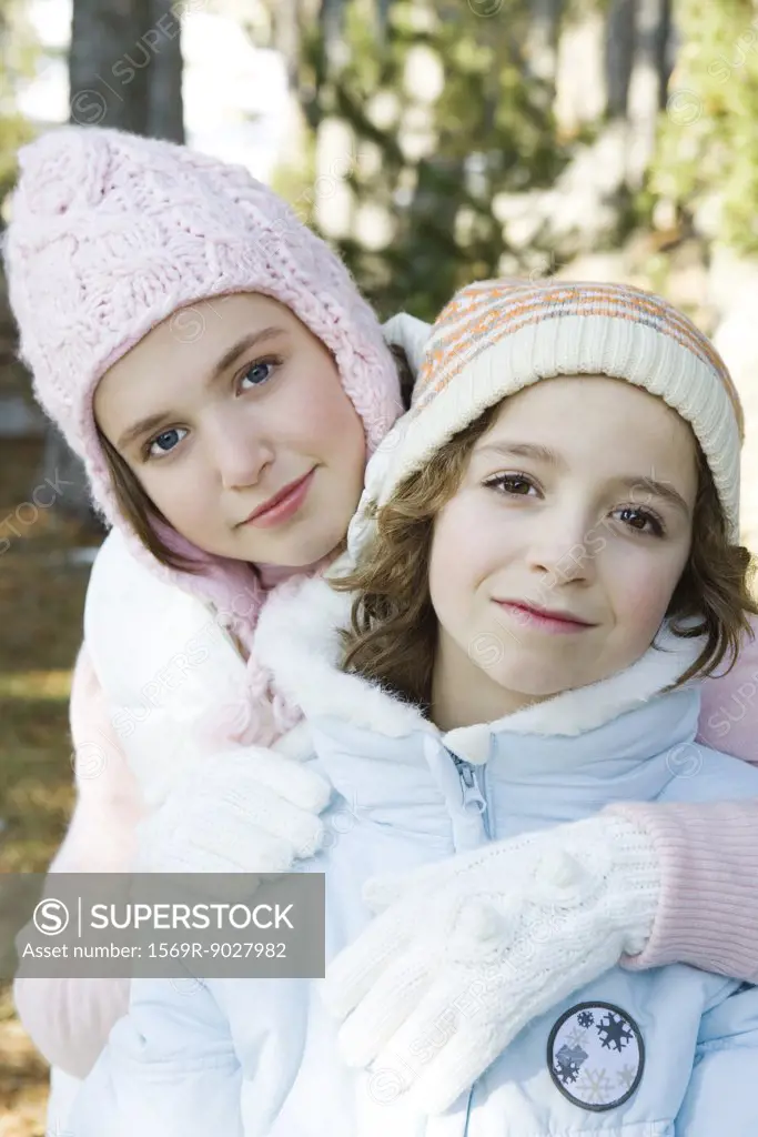 Two young friends smiling at camera, wearing knit hats, one's arm around the other's shoulder, portrait