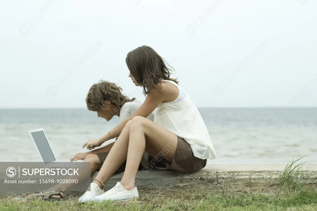 Teen girl and little brother sitting on the ground, using laptop computer, one pointing and looking over the other's shoulder