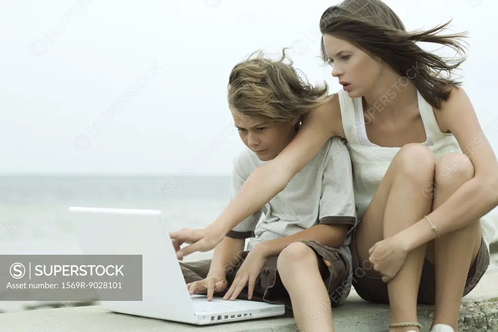 Teen girl and little brother using laptop computer together, girl pointing over boy's shoulder