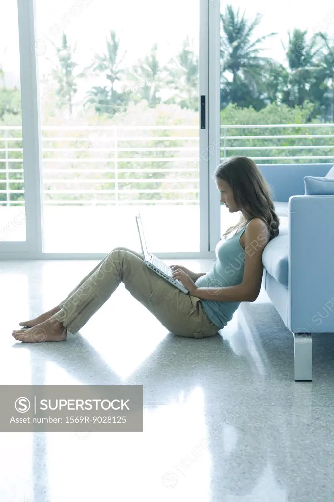 Teen girl sitting on the ground, leaning against sofa, using laptop computer