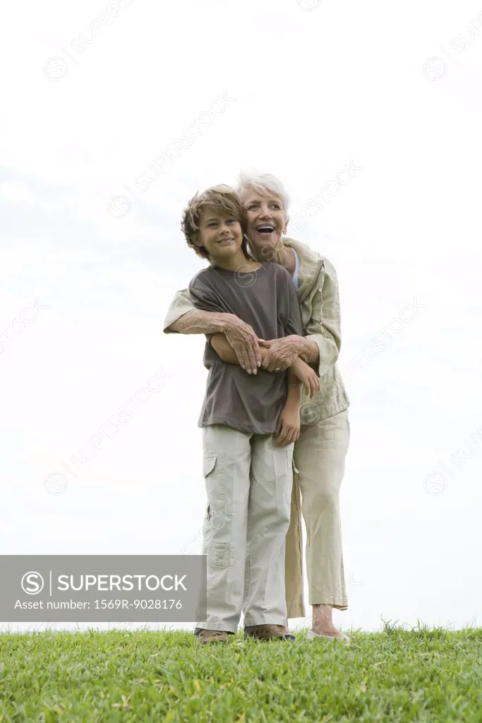 Senior woman and grandson standing on grass, woman hugging boy from behind, both smiling and looking out of frame, full length