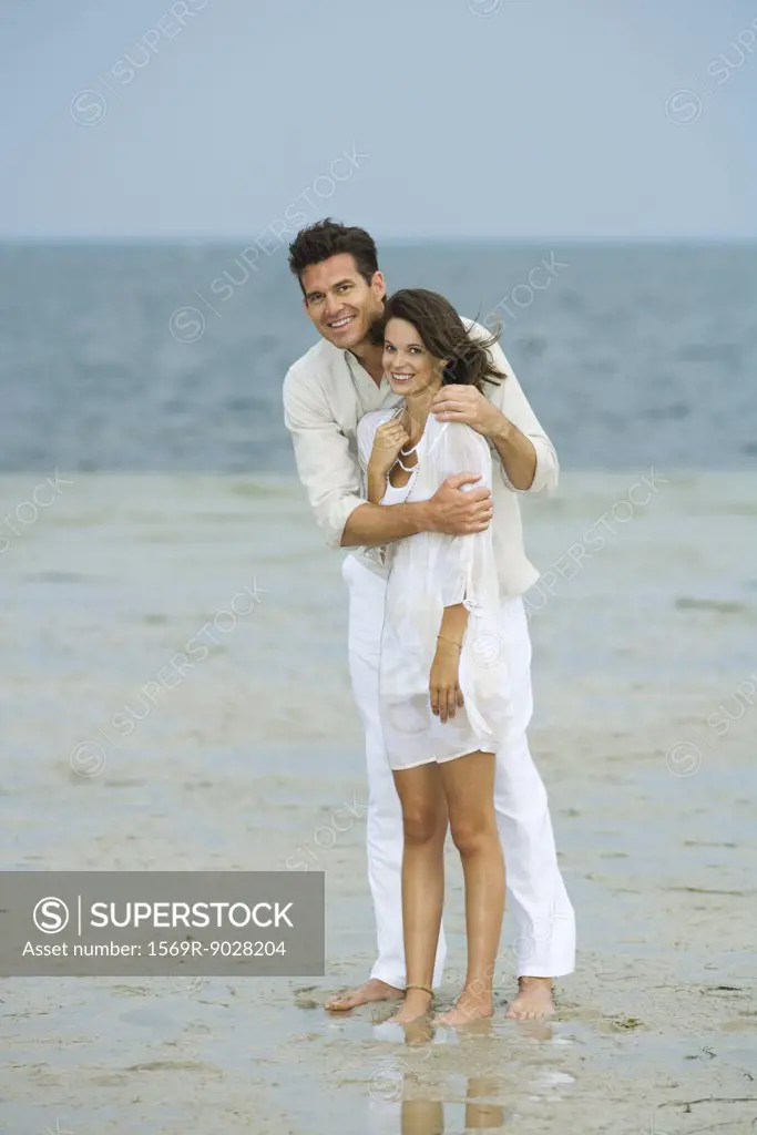 Man and young female companion on beach, standing together, looking at camera, full length