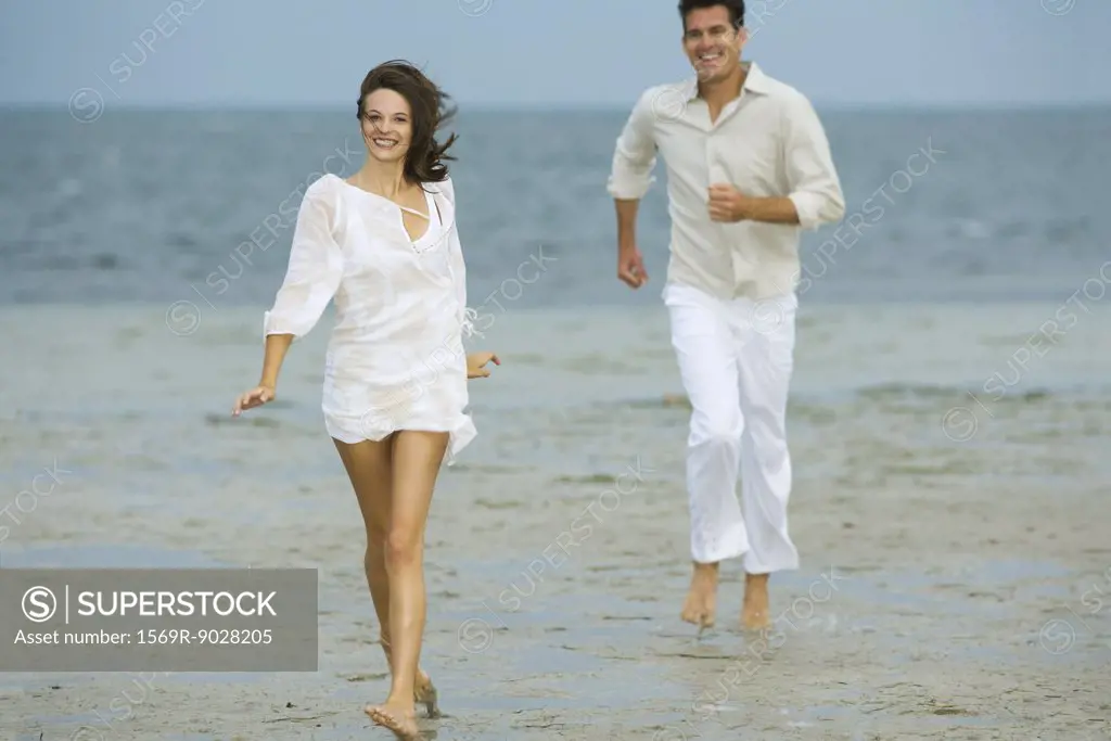 Man and young female companion on beach, hurrying toward camera, smiling, man in background, full length