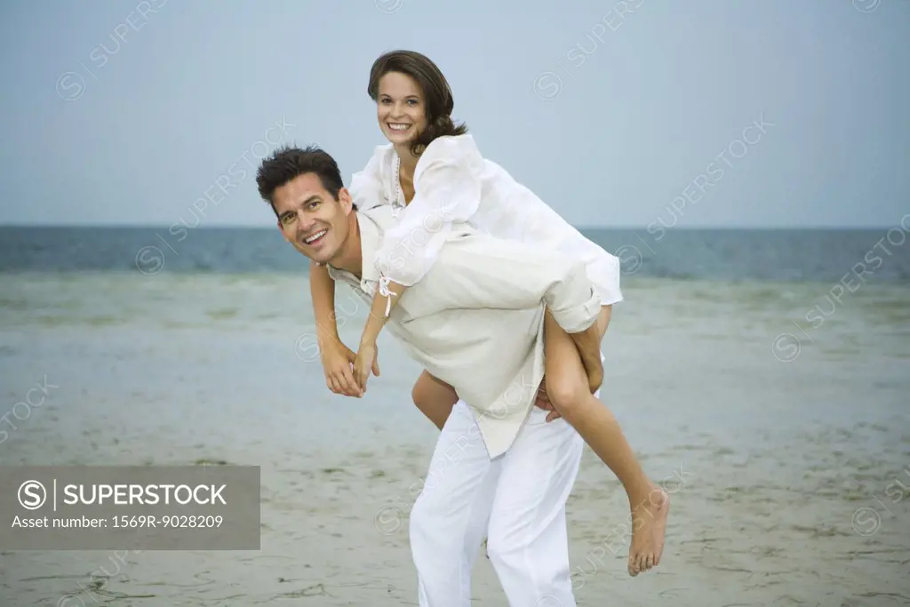 Man carrying young female companion piggyback on beach, smiling at camera