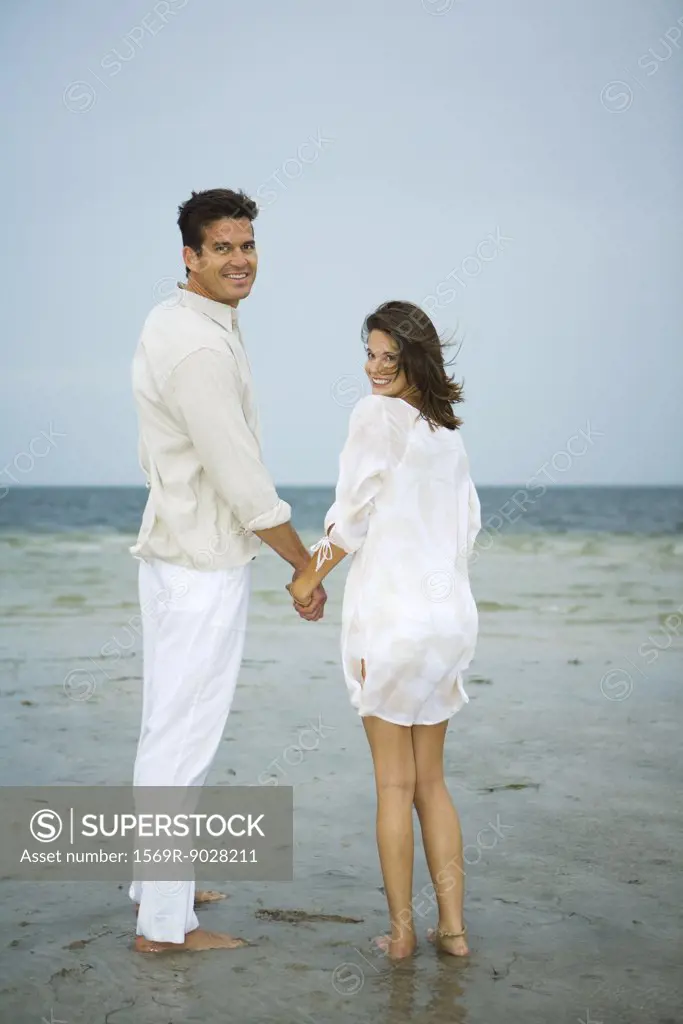 Man and young female companion on beach, holding hands, looking over shoulders at camera, full length