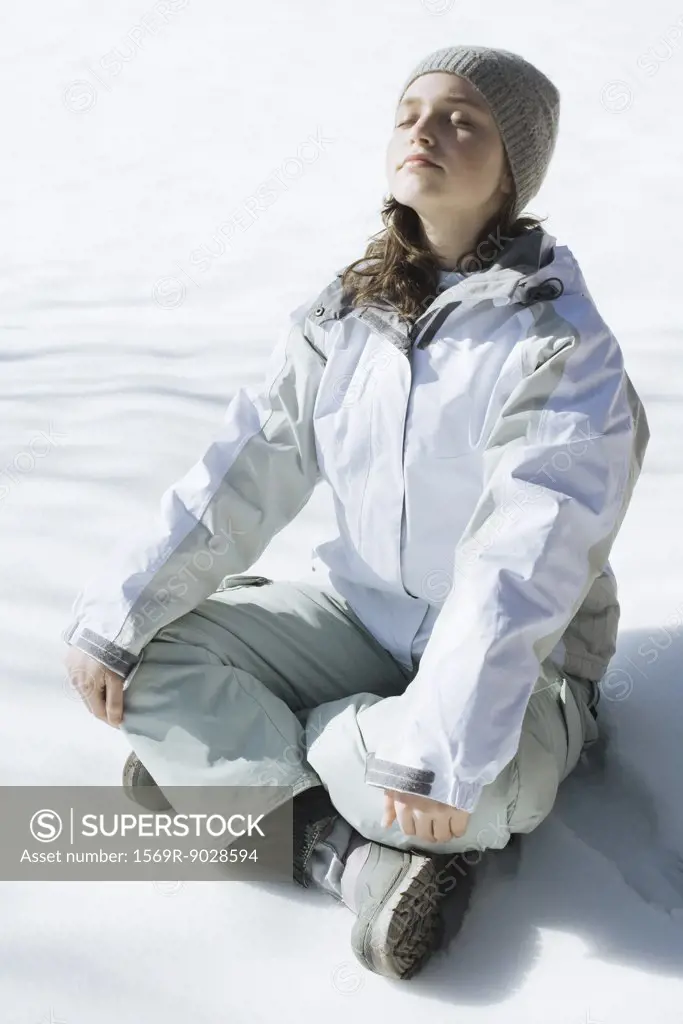 Teenage girl sitting indian style on snow with eyes closed and head back, full length