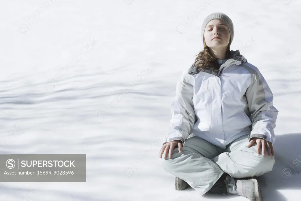 Teenage girl sitting indian style on snow with eyes closed and head back, full length