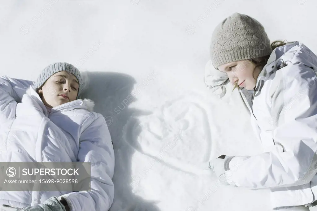 Teenage girls lying on snow, one drawing heart with initials in snow