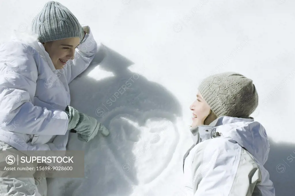 Teenage girls lying on snow, laughing, heart with initials drawn on surface of snow