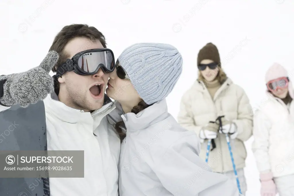 Young man standing with snowboard, giving thumbs up sign as teen girl kisses his cheek