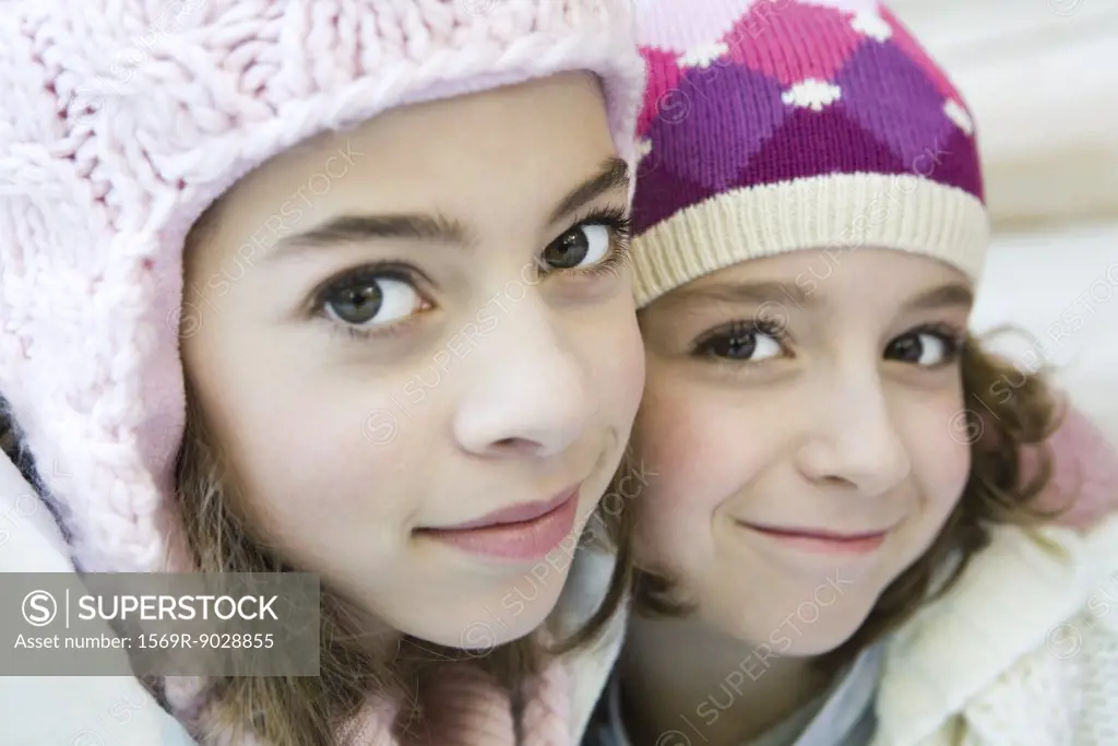 Two young sisters smiling at camera, both wearing knit hats, cheek to cheek, portrait