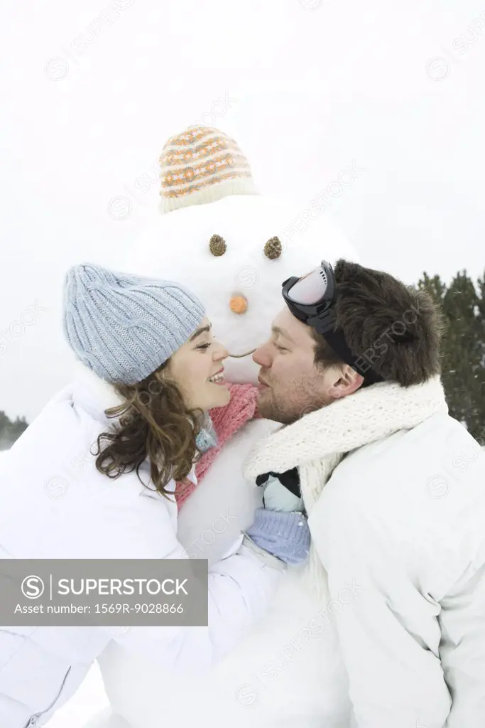 Young couple leaning towards each other in front of snowman, smiling, portrait