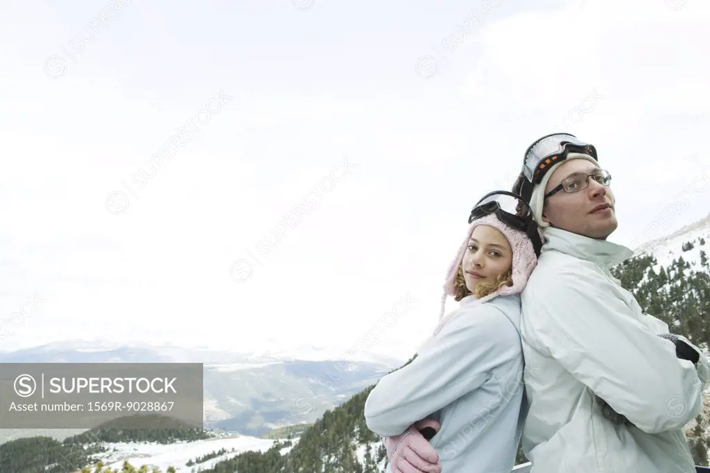 Young man and teen girl skiers standing back to back, smiling, girl looking at camera, portrait