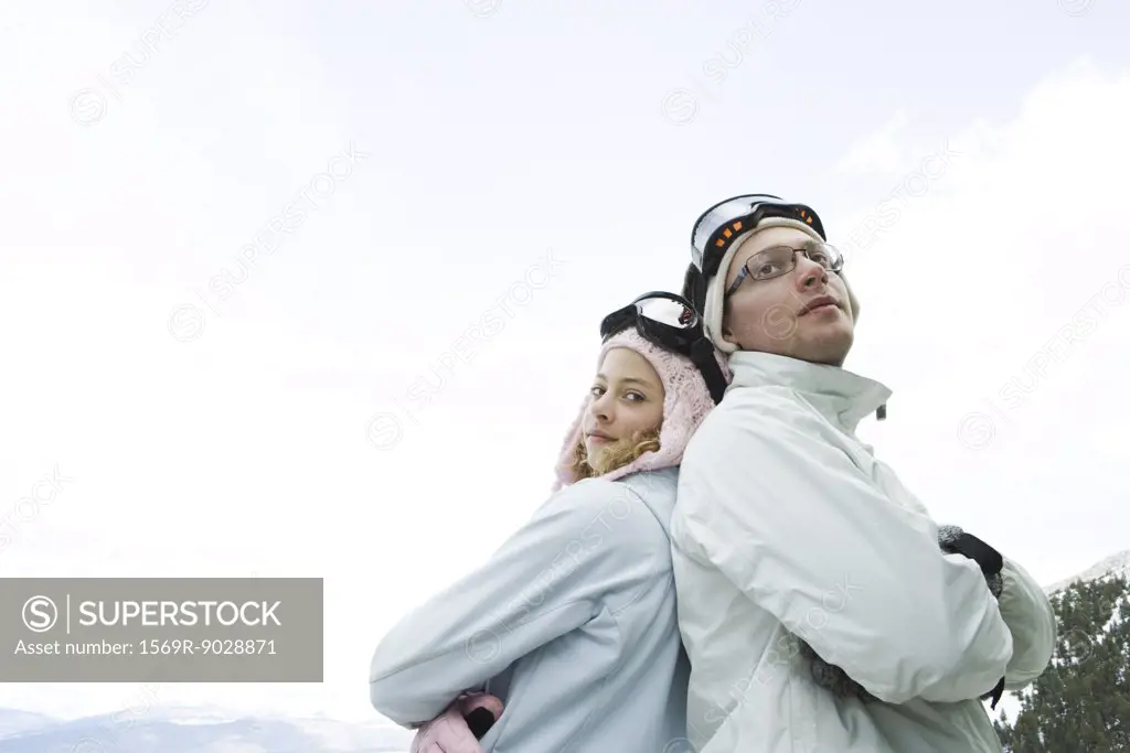 Teen girl and young man in ski gear, standing back to back, arms folded, smiling, girl looking at camera