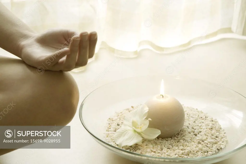 Female sitting in lotus position next to candle, cropped view of hand resting on bare knee
