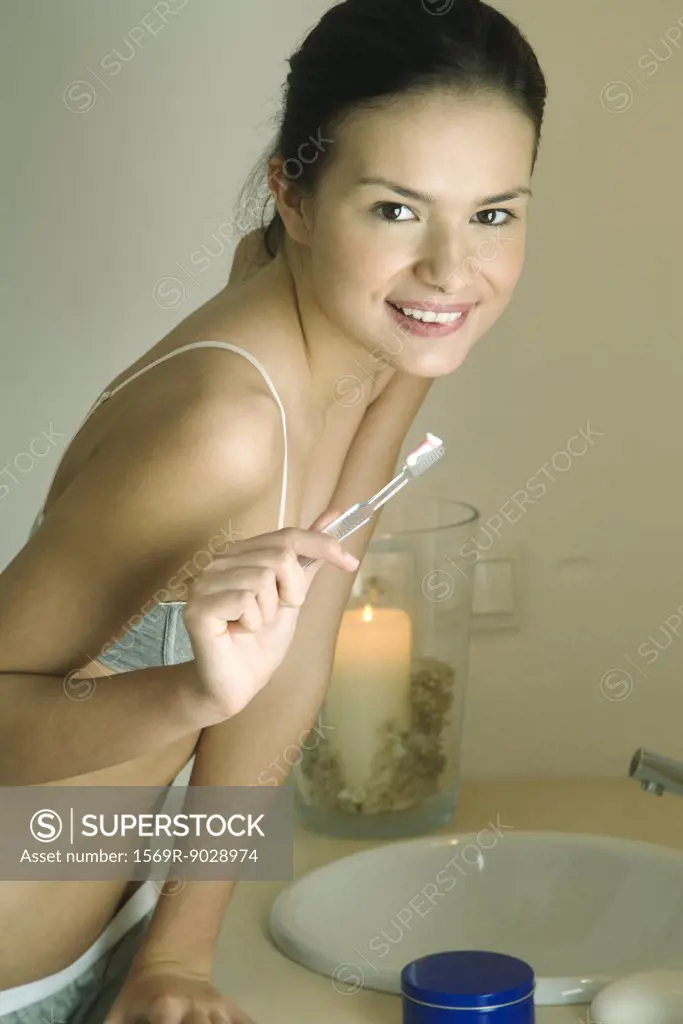 Young woman standing by sink, holding toothbrush, smiling at camera