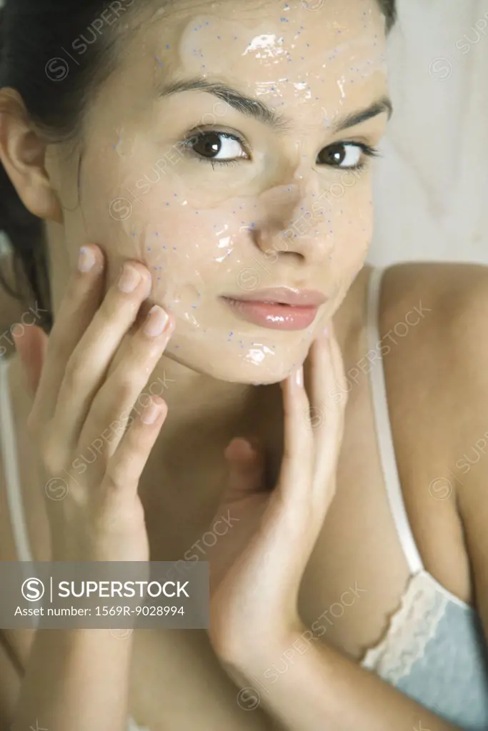 Young woman putting exfoliant mask on face, smiling at camera