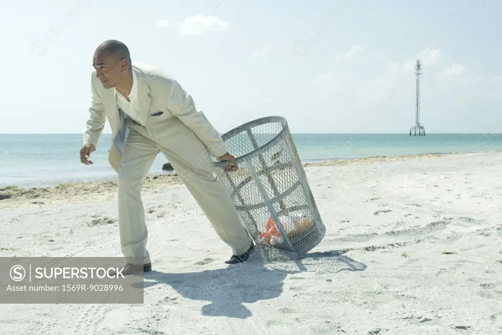 Man in suit pulling garbage can across sunny beach, full length