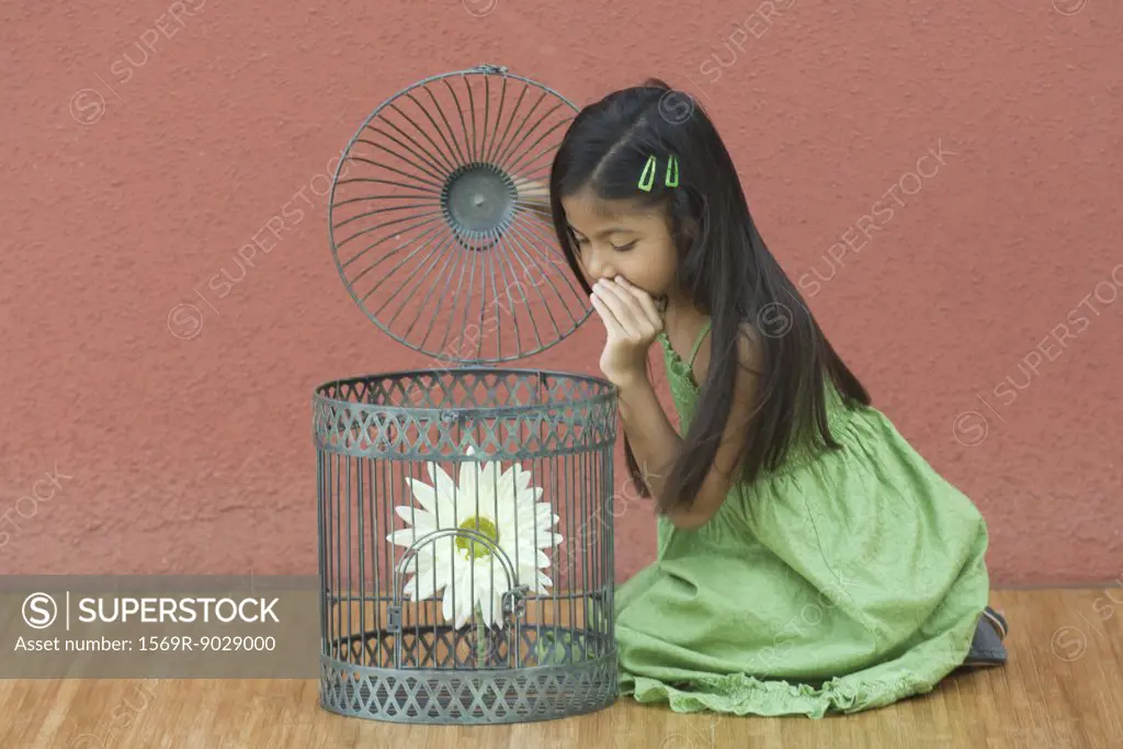 Girl kneeling on floor, whispering to flower in through open top of birdcage
