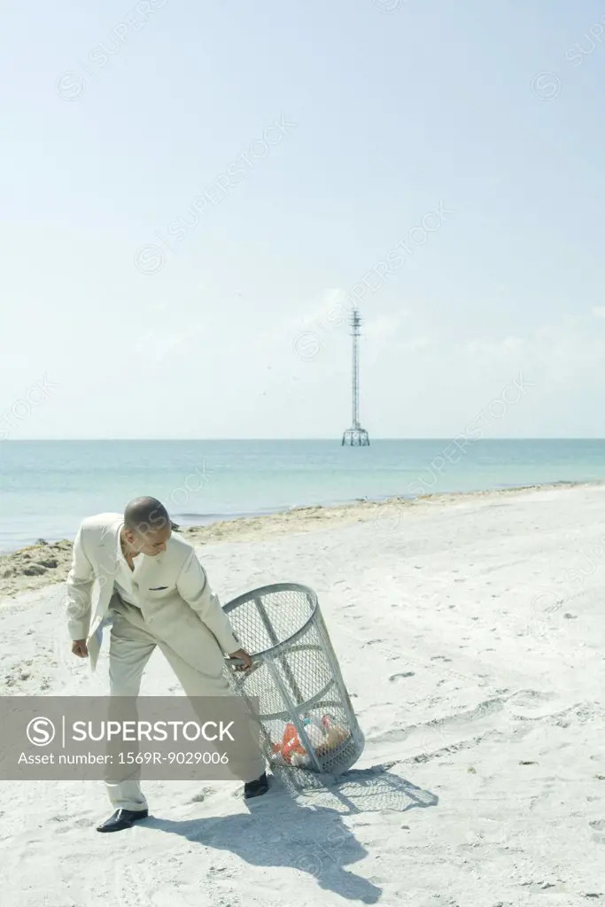 Man in suit pulling garbage can across sunny beach, looking back over shoulder, full length