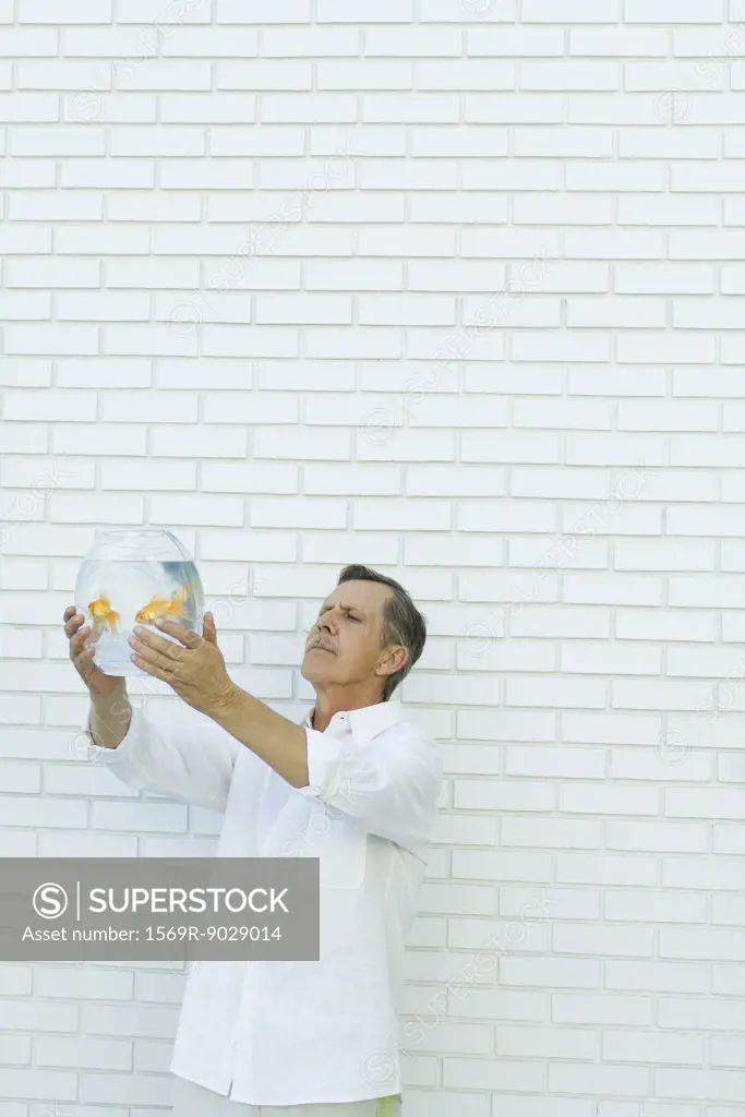 Man holding up goldfish bowl, standing in front of white brick wall, waist up
