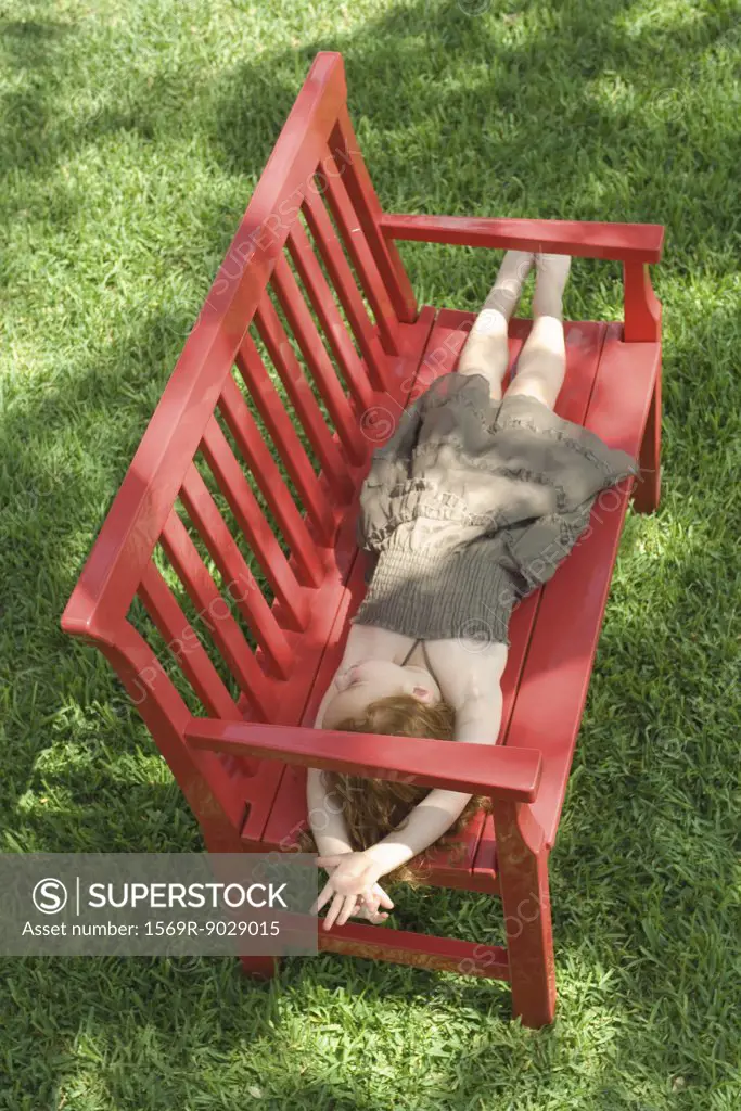 Little girl lying on red bench in shade, green grass in background, high angle view, full length
