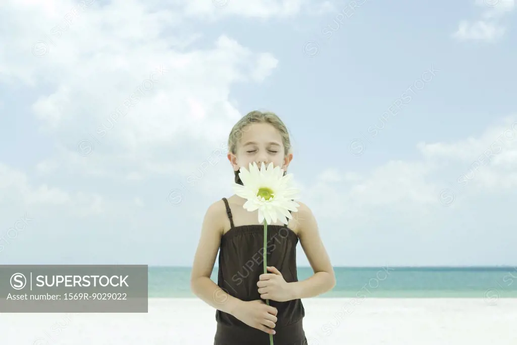 Girl holding up flower, eyes closed, standing on beach, waist up