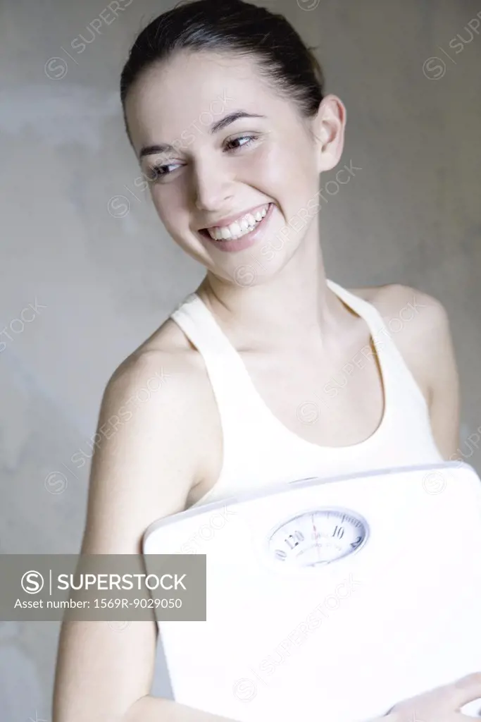 Young woman holding bathroom scale, smiling, portrait