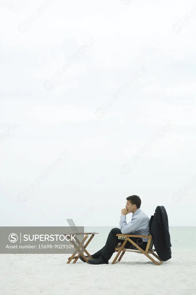 Businessman sitting in chair on beach, looking at laptop computer, side view