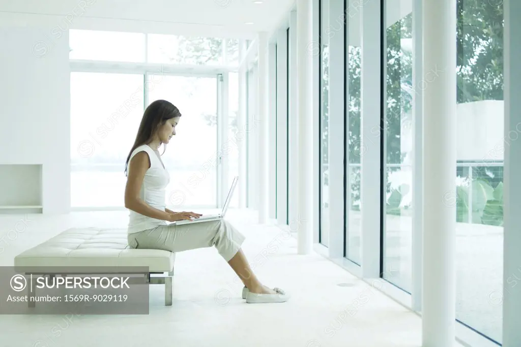 Young woman sitting on bench, using laptop computer, full length