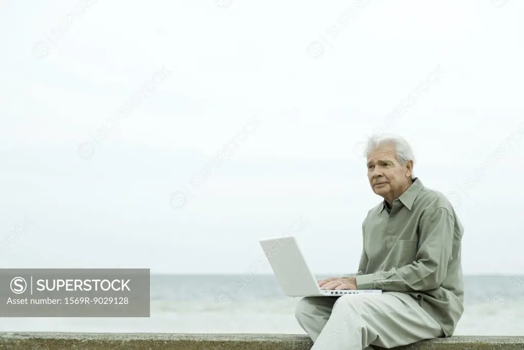 Senior man sitting near water's edge, using laptop