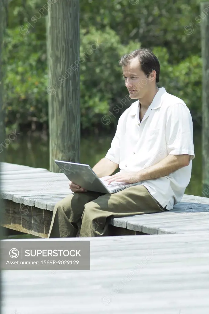 Man sitting on dock, using laptop computer, looking away