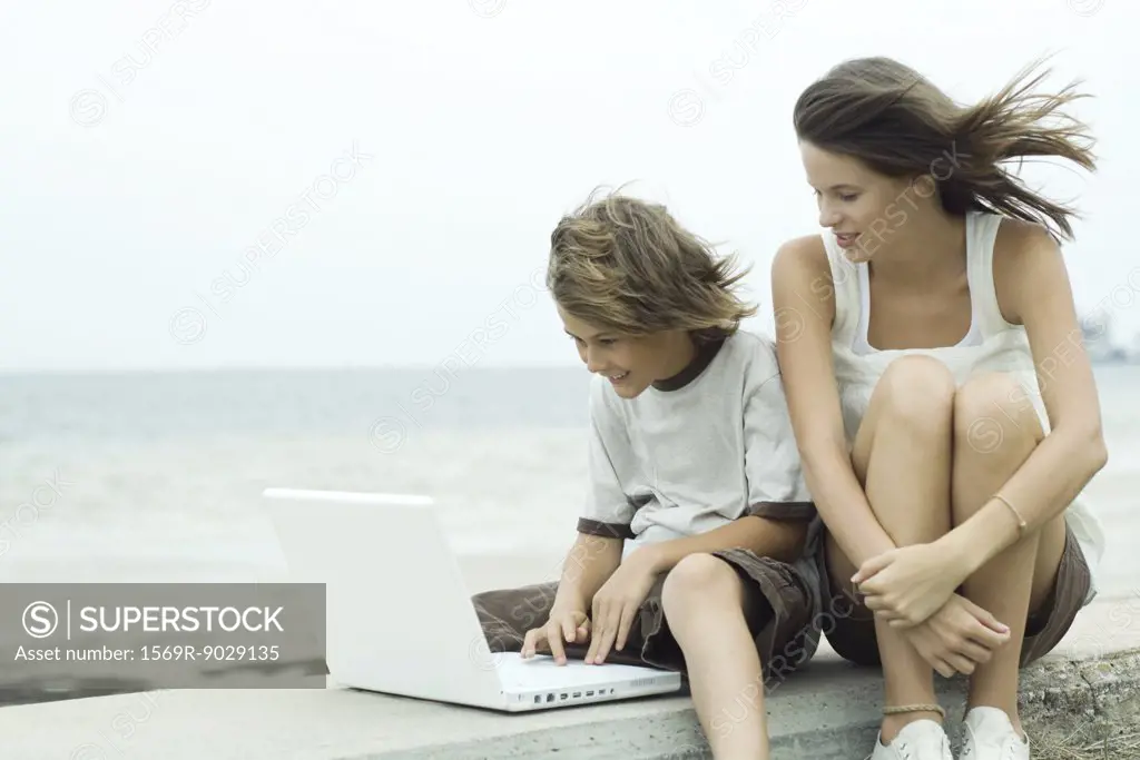 Teen girl and little brother using laptop near coast